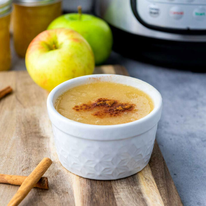 A small white ramekin filled with smooth, thick homemade applesauce, dusted with a stripe of cinnamon, placed on a wooden cutting board with cinnamon sticks. Two fresh apples and an Instant Pot are visible in the soft-focus background.