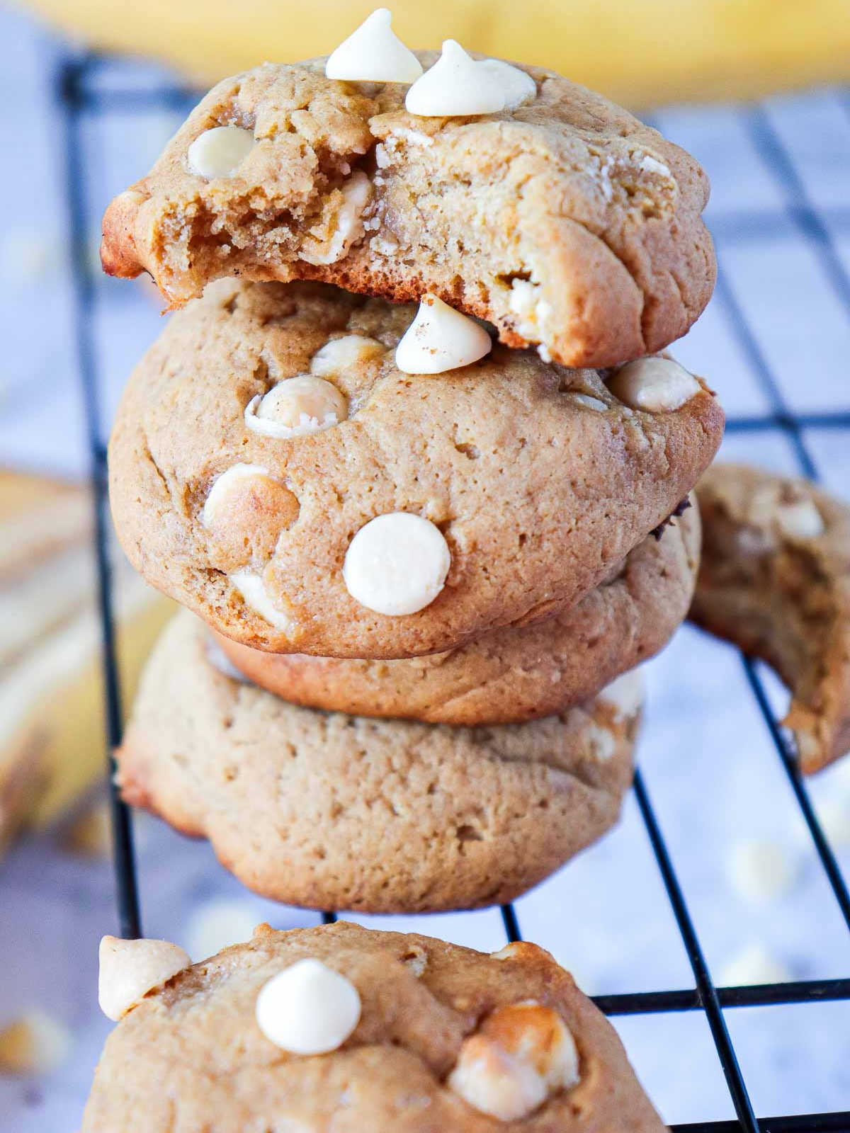 A close-up stack of four soft-baked banana pudding cookies on a black wire cooling rack, studded with white chocolate chips. The top cookie has a bite taken out of it, revealing a moist interior.