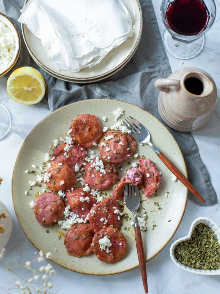 An overhead view of round, pink beet ravioli stuffed with goat cheese, plated on a beige ceramic dish and topped with crumbled feta cheese and chopped chives. A fork is cutting one open to show the bright filling. The dish is surrounded by a glass of red wine, a lemon half, and a small heart-shaped bowl of dried herbs.
