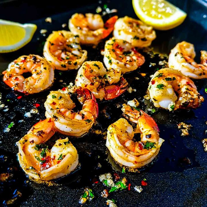 A close-up of several peeled, pink shrimp cooking on a black griddle or flat-top grill, glistening with melted garlic butter, red chili flakes, and chopped herbs. Two lemon wedges are visible in the background.