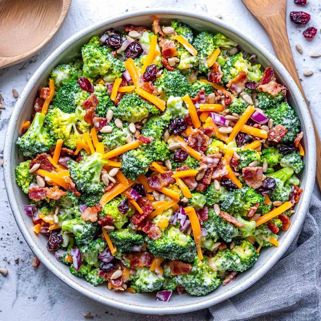 An overhead shot of a large bowl of creamy broccoli salad, loaded with toppings including crispy bacon, shredded cheddar cheese, dried cranberries, and sunflower seeds.