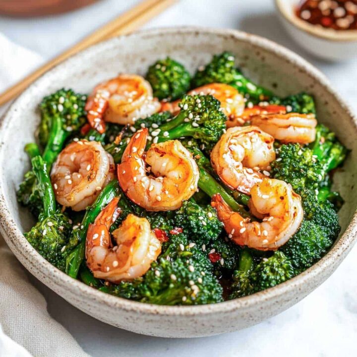 A close-up view of a bowl of glossy shrimp and broccolini stir-fry, seasoned with red chili flakes and sesame seeds. Chopsticks and a small bowl of sauce are visible in the soft-focus background.