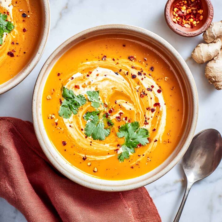 An overhead view of a bowl of vibrant orange carrot ginger soup, made creamy with cottage cheese, garnished with a swirl of cream, fresh cilantro, and red chili flakes. A silver spoon and a piece of fresh ginger are visible on the side.