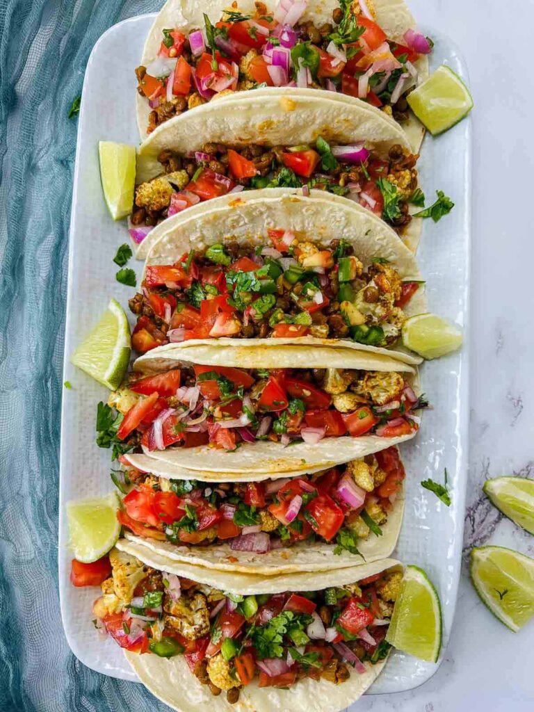 An overhead shot of a long white platter lined with cauliflower and lentil tacos, topped with fresh pico de gallo and served with lime wedges.