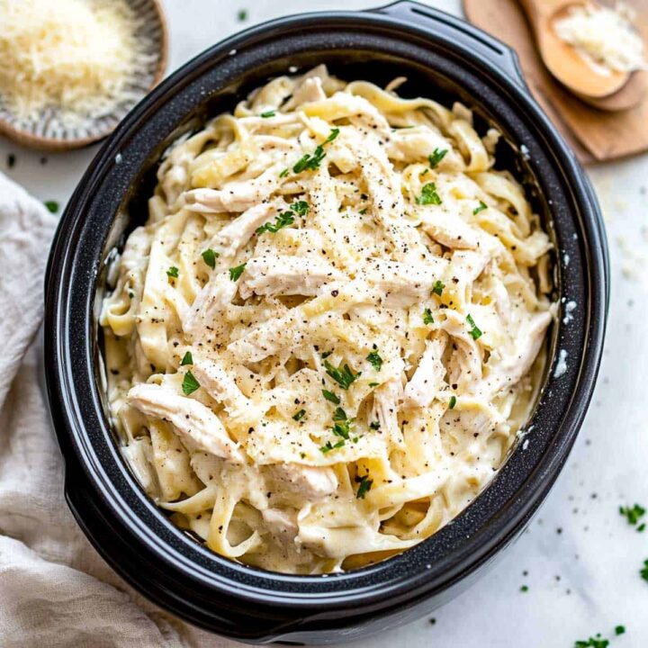 An overhead view of creamy slow cooker chicken alfredo, featuring fettuccine pasta and shredded chicken coated in a white sauce, garnished with grated Parmesan cheese, black pepper, and chopped parsley, served inside a black oval slow cooker insert.