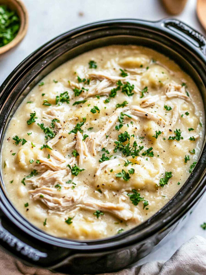 An overhead view of creamy slow cooker chicken and dumplings inside a black oval slow cooker insert, filled with a thick, savory broth, fluffy dumplings, and shredded chicken, garnished generously with fresh chopped parsley and black pepper.