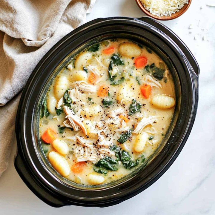 An overhead view of creamy chicken and gnocchi soup served inside a black oval slow cooker insert, filled with plump potato gnocchi, shredded chicken, chopped carrots, and spinach, garnished with cracked black pepper and a sprinkle of Parmesan cheese.
