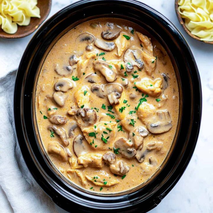 An overhead view of creamy slow cooker chicken stroganoff, featuring chicken chunks and sliced mushrooms in a rich, tan sauce, garnished with chopped fresh parsley, served inside a black oval slow cooker insert. Small bowls of egg noodles are visible in the upper corners.