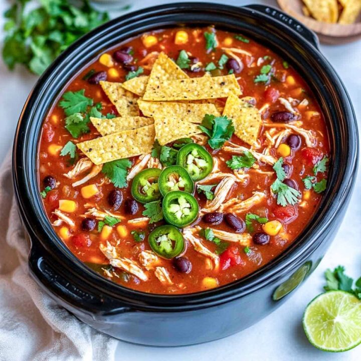 An overhead view of a black slow cooker or pot filled with vibrant red chicken tortilla soup, loaded with shredded chicken, black beans, corn, and diced tomatoes, garnished with tortilla chips, sliced jalapeños, and fresh cilantro.