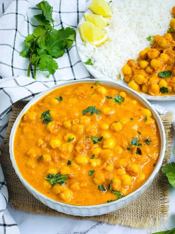 An overhead close-up of a white bowl filled with creamy orange chickpea and mango curry, garnished with fresh cilantro, sitting on a burlap mat. A side plate with white rice, lemon wedges, and a small serving of the curry is visible in the background.
