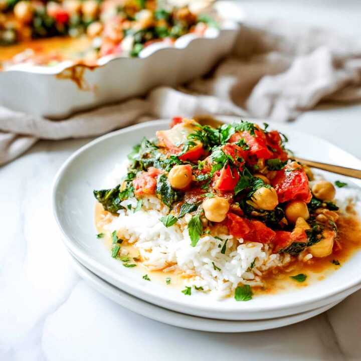 A close-up of a white plate with a serving of chickpea, spinach, and tomato casserole ladled over a bed of fluffy white rice, with the main baking dish in the background.
