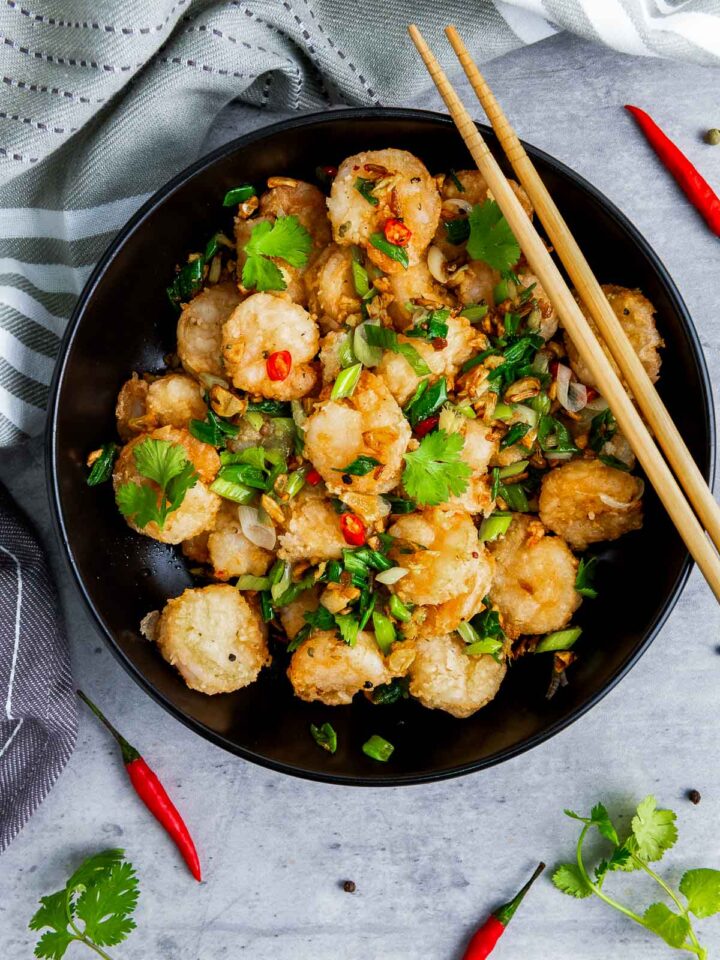 An overhead view of a black bowl filled with crispy, battered Chinese salt and pepper shrimp, tossed with chopped green onions, fried garlic, red chili flakes, and cilantro leaves. A pair of wooden chopsticks rests on top of the dish, with fresh chili peppers scattered around the bowl.