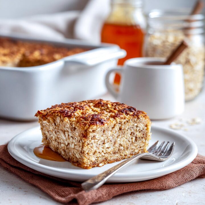 A close-up of a square slice of cinnamon baked oatmeal on a plate with a fork, with the full baking dish visible in the background.