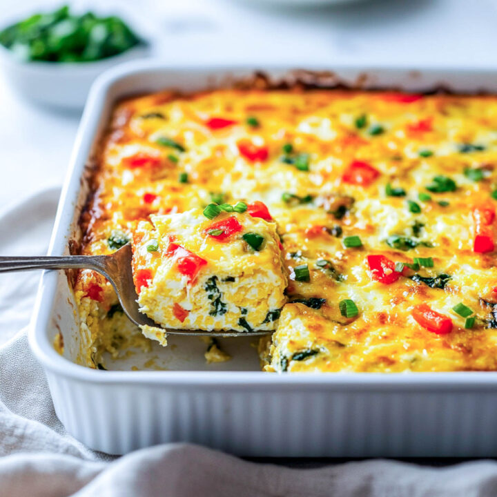 A slice of a fluffy egg casserole, filled with red peppers and spinach, is being lifted from a white baking dish with a spatula.