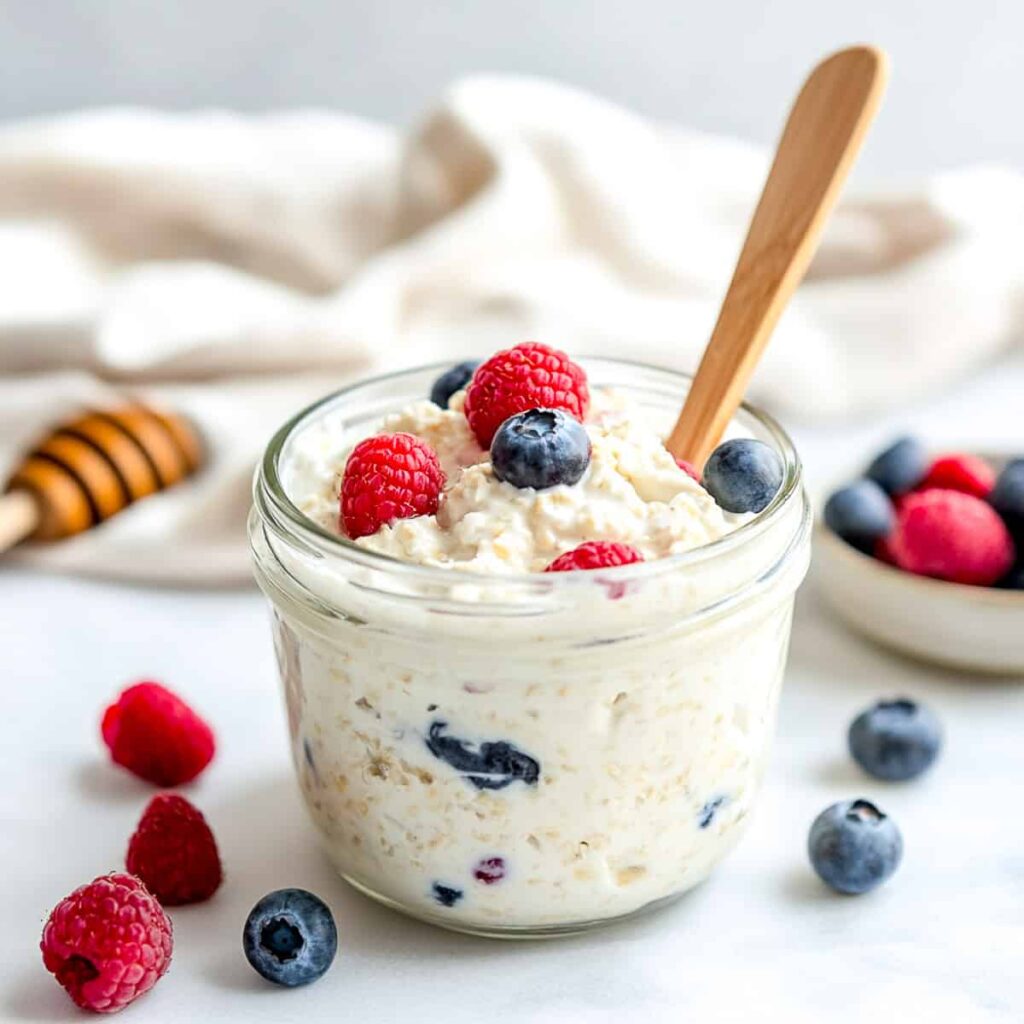 A close-up of a glass jar of overnight oats topped with fresh raspberries and blueberries, with a wooden spoon in the jar and a honey dipper in the background.