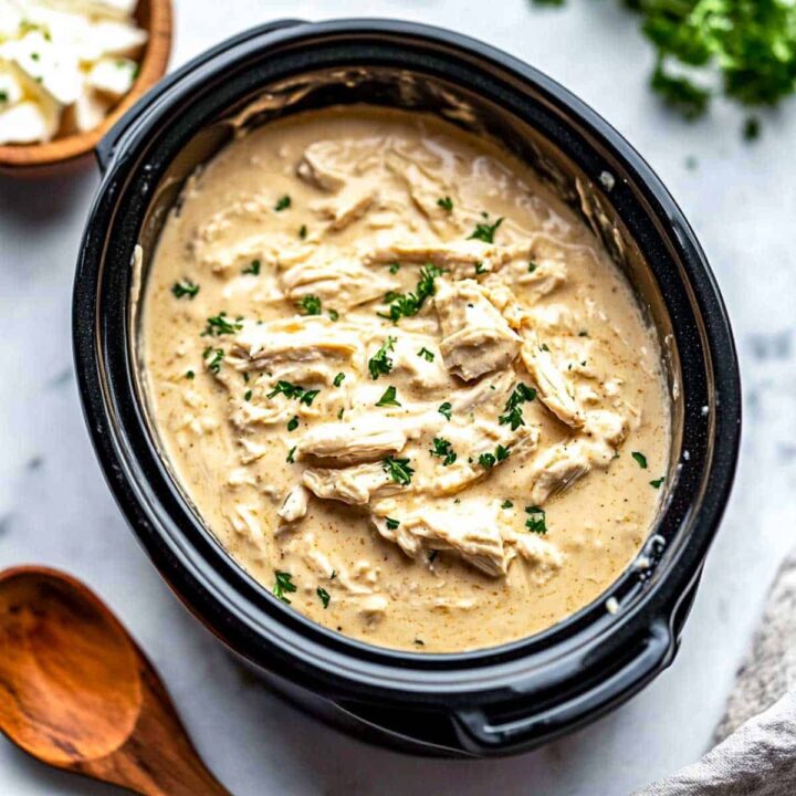 An overhead view of creamy slow cooker shredded chicken, likely made with cream cheese and a golden sauce, garnished with chopped fresh parsley, served inside a black oval slow cooker insert. A wooden spoon and a bowl of toppings (possibly cheese) are partially visible.