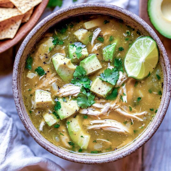 An overhead view of a bowl of chicken avocado soup in a light broth, filled with shredded chicken and topped generously with cubed avocado, cilantro leaves, cracked black pepper, and a wedge of lime. A bowl of tortilla chips is partially visible in the upper left corner.