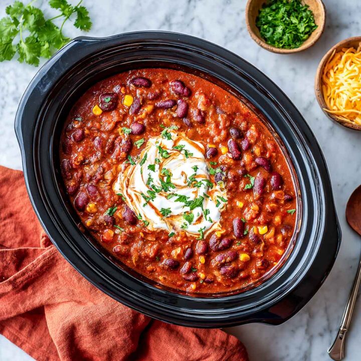 An overhead view of hearty slow cooker chili (likely made high-protein with cottage cheese), filled with kidney beans and corn in a rich red broth, topped with a swirl of sour cream or yogurt and fresh parsley. Bowls of shredded cheese and chopped cilantro are visible on the side.