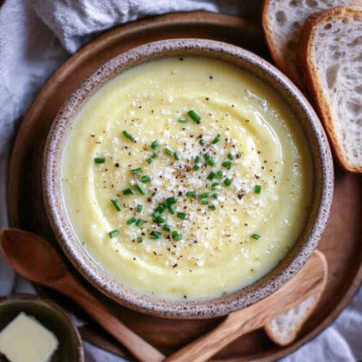 An overhead view of a bowl of thick, creamy potato leek soup, garnished with grated Parmesan cheese, freshly cracked black pepper, and chopped chives. The bowl is served on a wooden plate next to slices of crusty bread and a small dish of butter.