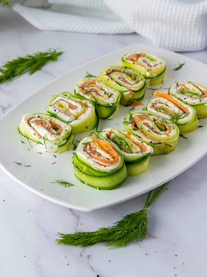 A rectangular white platter holds twelve servings of cucumber smoked salmon and cream cheese roll-ups. The rolls are made with thin slices of cucumber wrapped around cream cheese and slices of smoked salmon, and are garnished with fresh dill and everything bagel seasoning.