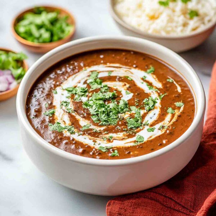 A bowl of rich, dark brown Dal Makhani (creamy lentil dish) garnished with a swirl of fresh cream and chopped cilantro. Bowls of white rice, chopped red onion, and cilantro are visible in the background.