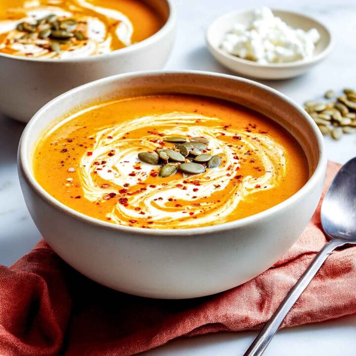 A bowl of vibrant orange, creamy pumpkin soup, garnished with a swirl of cream, toasted pepitas (pumpkin seeds), and red chili flakes. A spoon and a small bowl of cottage cheese are visible in the background.