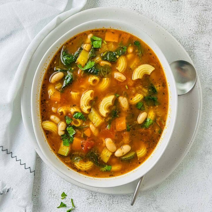 An overhead view of a white bowl filled with a hearty minestrone soup, featuring a tomato-based broth with elbow pasta, cannellini beans, zucchini, carrots, celery, and dark leafy greens, garnished with fresh basil or parsley.