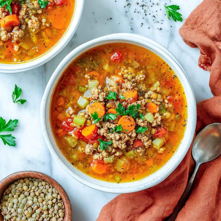 An overhead view of a white bowl filled with a chunky, orange-broth turkey and lentil soup, loaded with ground meat, carrots, celery, and diced tomatoes, garnished with fresh parsley and black pepper. A bowl of uncooked lentils and a silver spoon are visible on the marble surface.