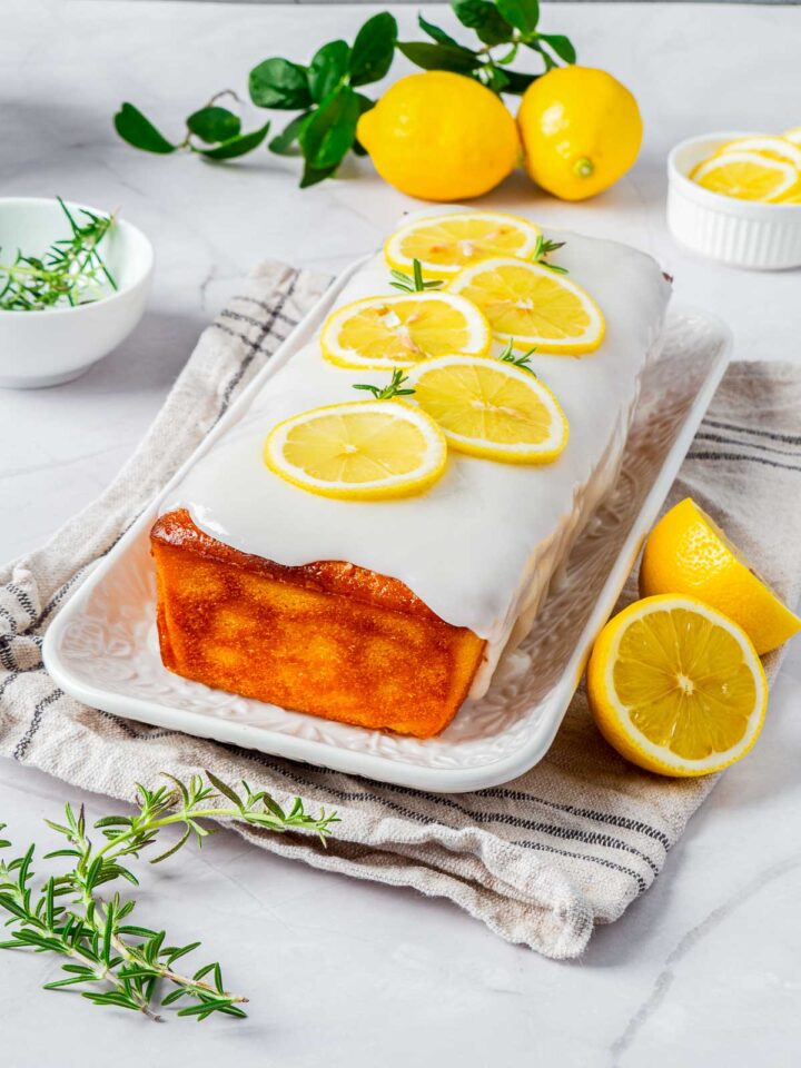 A golden brown lemon loaf cake topped with a thick white glaze and garnished with thin, overlapping slices of fresh lemon and small sprigs of rosemary. It sits on a white platter on a marble surface with whole lemons and rosemary sprigs scattered around it.