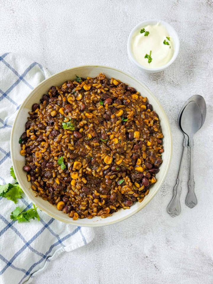 An overhead close-up of a bowl filled with Mexican rice and beans, featuring a rich, dark mixture of rice, black beans, corn, and spices, garnished with chopped cilantro. A small bowl of sour cream and two silver spoons are on the right.