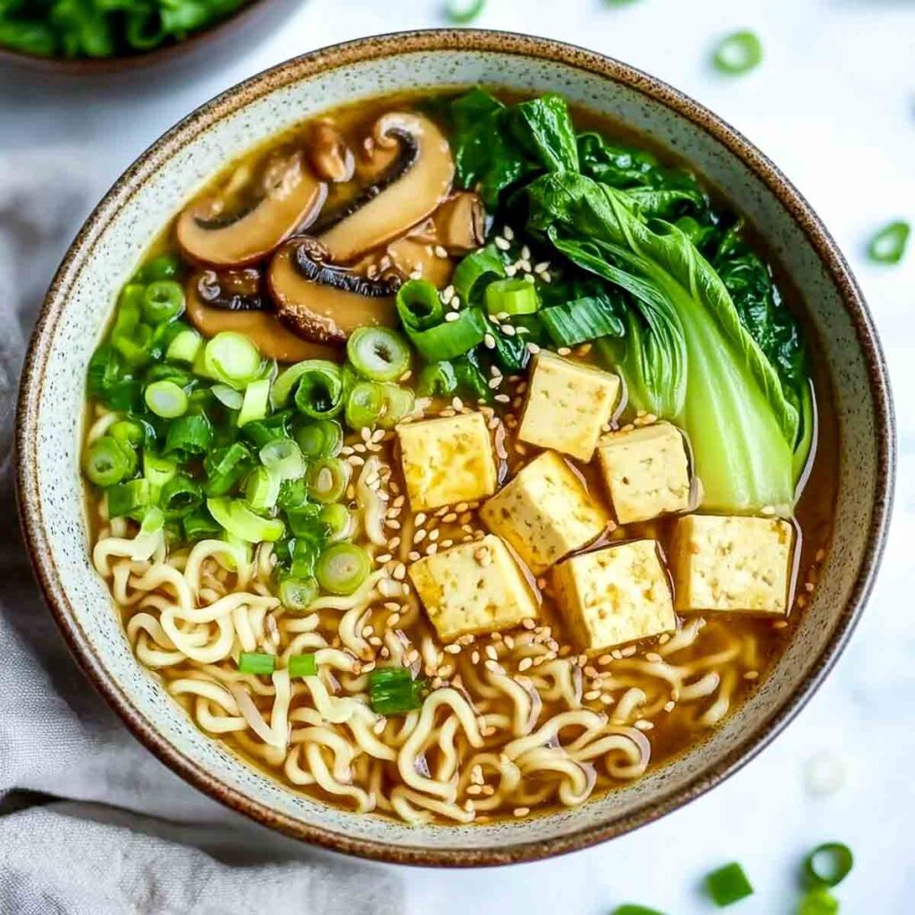An overhead shot of a rustic bowl of miso ramen, artfully topped with cubes of tofu, sliced mushrooms, baby bok choy, and a generous amount of chopped green onions.