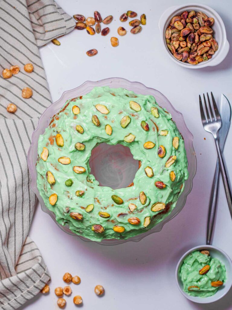 An overhead shot of a pistachio bundt cake covered in a thick, light green glaze and topped with chopped pistachio pieces, sitting on a glass cake stand. A small bowl of shelled pistachios and a bowl of extra glaze are visible nearby.