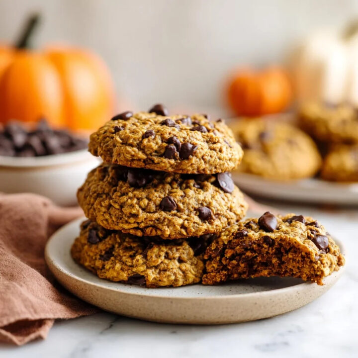 A close-up of a stack of pumpkin oatmeal chocolate chip cookies on a plate, with one broken to show the interior, and small pumpkins in the background.
