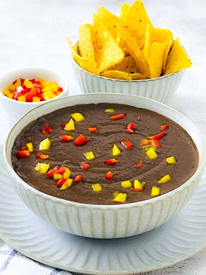 A bowl of thick, smooth, dark refried black beans topped with diced red and yellow bell peppers, served with a large bowl of yellow tortilla chips in the background.