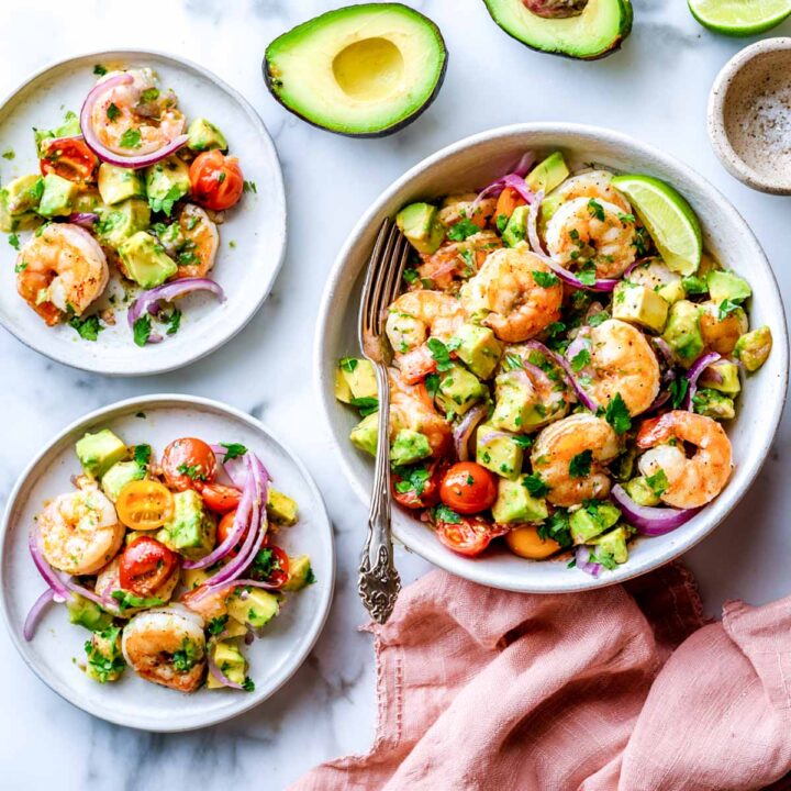 An overhead view of a large bowl and two small side plates filled with a shrimp and avocado salad, mixed with halved cherry tomatoes and sliced red onion, and garnished with cilantro and a lime wedge. A halved avocado is visible in the background.