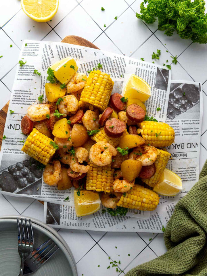 A classic shrimp boil spread piled high on a sheet of newspaper, featuring seasoned shrimp, sliced smoked sausage, corn on the cob sections, and small golden potatoes, garnished with lemon wedges and fresh parsley.