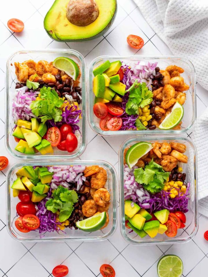 An overhead view of four glass meal prep containers filled with shrimp taco rice bowls, featuring layers of seasoned shrimp, black beans, corn, red cabbage, cherry tomatoes, avocado chunks, and red onion, garnished with cilantro and a lime wedge. A halved avocado is visible above the containers.