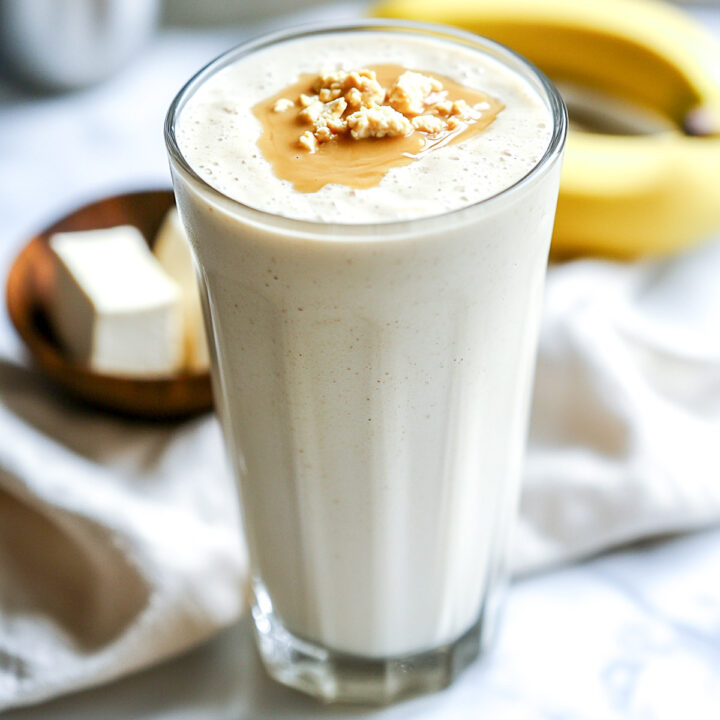 A close-up of a tall glass of a creamy protein shake, topped with a dollop of peanut butter, with bananas and a block of silken tofu in the background.