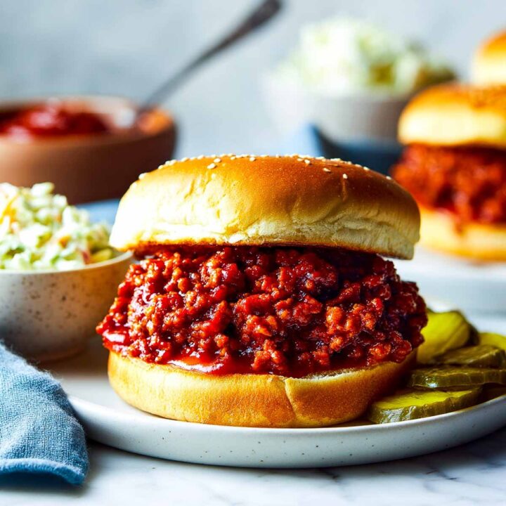 A classic Sloppy Joe sandwich featuring a thick, saucy pile of seasoned ground meat filling on a toasted, sesame seed bun, served on a white plate with dill pickle slices. Bowls of coleslaw and a small cup of extra sauce are visible in the soft-focus background.