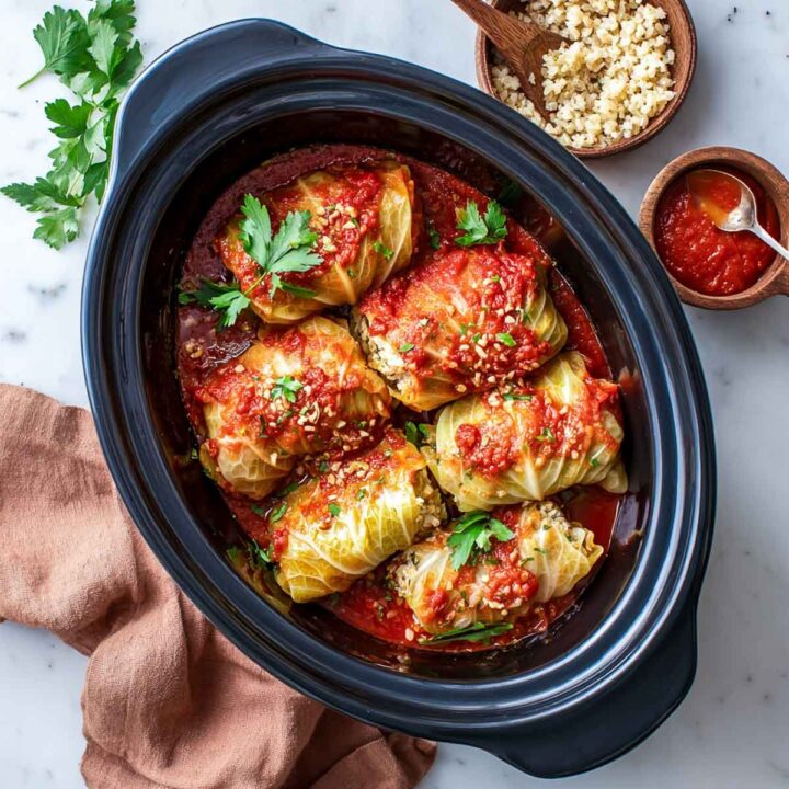 An overhead view of six stuffed cabbage rolls simmering in a red tomato sauce inside a black oval slow cooker insert. The cabbage rolls are filled with a mixture of cottage cheese and brown rice, topped with sauce, a sprinkle of garnish, and fresh parsley. Small bowls of sauce and cooked rice are visible in the top right.
