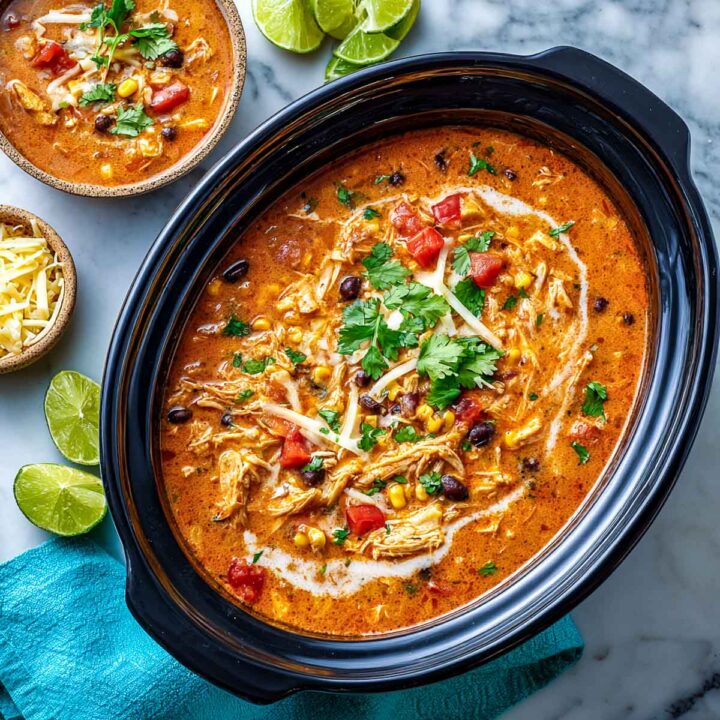 An overhead view of slow cooker chicken enchilada soup (likely made creamy with cottage cheese) in a black oval slow cooker insert, filled with shredded chicken, black beans, corn, and tomatoes. It is garnished with shredded cheese, a swirl of cream, and fresh cilantro. Lime wedges and a small bowl of shredded cheese are visible on the marble surface.
