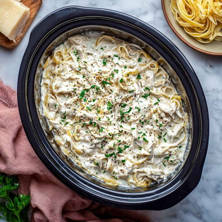 An overhead view of creamy slow cooker chicken alfredo (likely high-protein with cottage cheese) with fettuccine pasta and shredded chicken, garnished with black pepper and fresh parsley, served inside a black oval slow cooker insert. A block of Parmesan cheese and a plate of uncooked noodles are visible in the background.