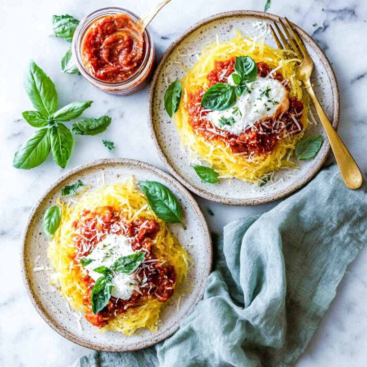 An overhead view of two plates of spaghetti squash "noodles," topped with a chunky red marinara sauce, a dollop of creamy cottage cheese, grated Parmesan, and fresh basil leaves. A small jar of the sauce and a sprig of basil are visible in the upper left corner.