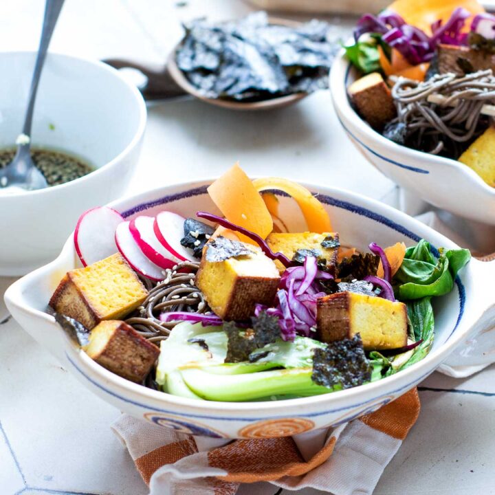 An overhead shot of two white bowls filled with a colorful smoked tofu poke bowl, containing soba noodles, carrot ribbons, red cabbage, radishes, and crumbled seaweed.