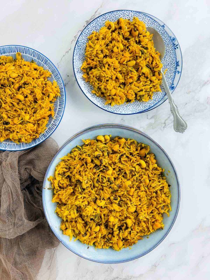 An overhead view of three bowls of golden yellow, spiced rice and black-eyed peas, or Hoppin' John, served on white and blue patterned plates on a marble surface.