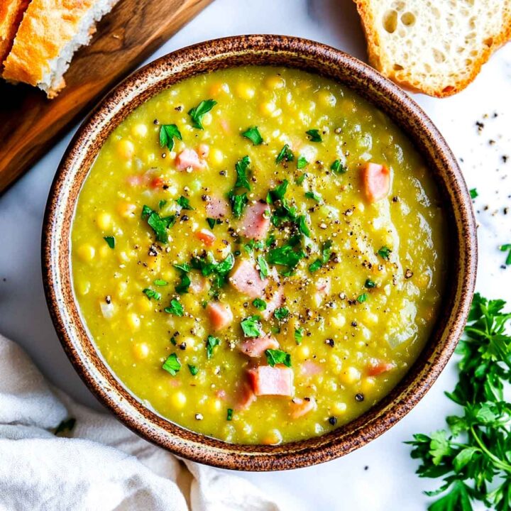 An overhead view of a bowl of thick, chunky yellow-green split pea and ham soup, garnished with diced ham chunks, fresh chopped parsley, and black pepper. Slices of crusty bread are visible on the side.