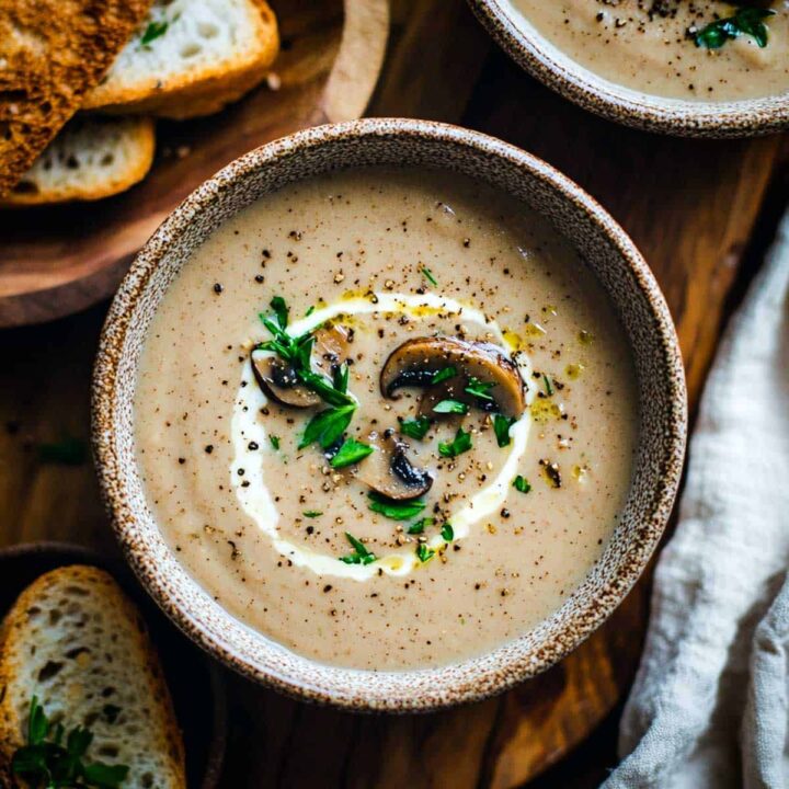 A bowl of thick, creamy mushroom soup, garnished with a swirl of cream, sliced sautéed mushrooms, freshly cracked black pepper, and chopped herbs. Toasted slices of bread are visible on the left.