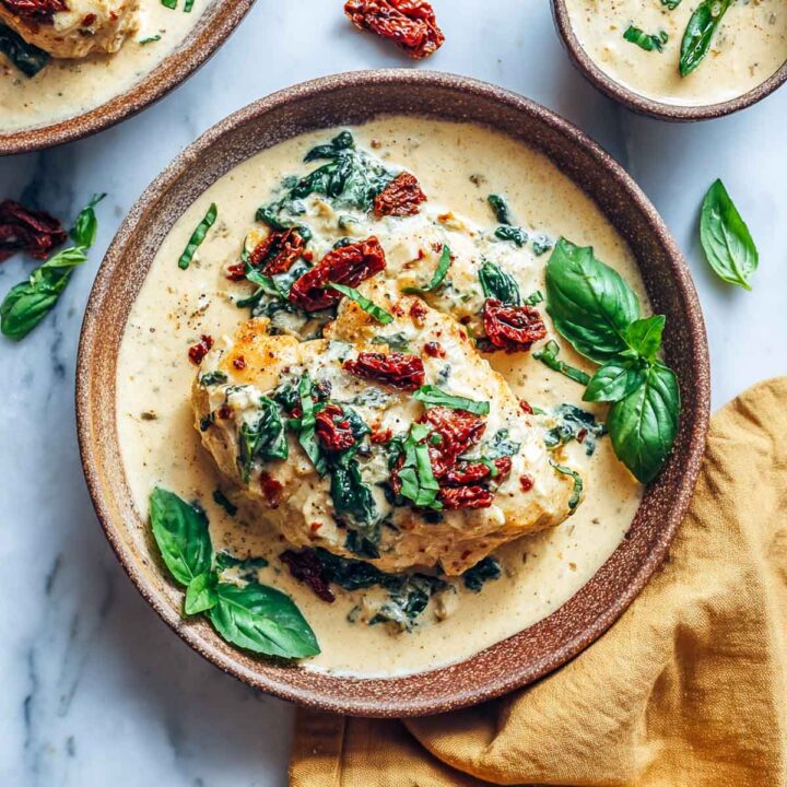 A portion of Tuscan chicken served in a creamy, golden sauce (made with cottage cheese), topped with wilted spinach, sun-dried tomatoes, and fresh basil leaves, plated in a rustic brown bowl on a marble surface.