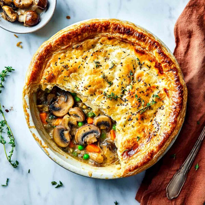 A deep-dish vegan mushroom pot pie in a ceramic dish, featuring a golden, flaky puff pastry crust with a slice removed to reveal a creamy filling of sliced mushrooms, carrots, and peas. The crust is garnished with thyme.