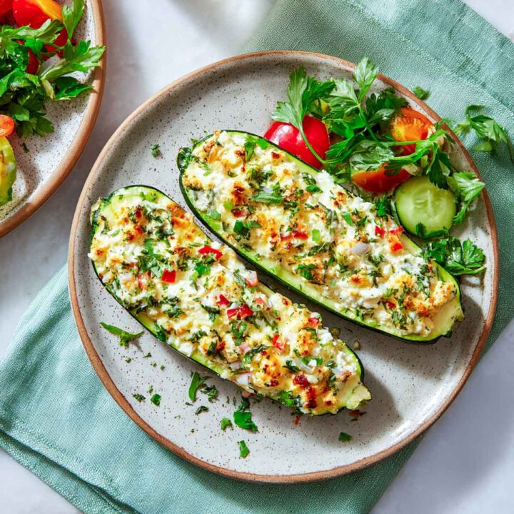 Two baked zucchini boats, stuffed with a cheesy white cottage cheese mixture, red bell peppers, and fresh herbs, served on a round plate with a side salad of chopped tomatoes, cucumber slices, and parsley.